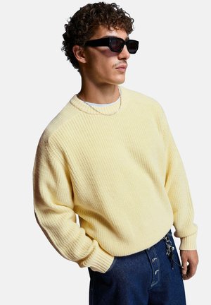 Light yellow ribbed knit sweater with a round neckline, worn over a white shirt, paired with dark blue jeans and black sunglasses.