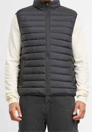 Mężczyzna ubrany w czarną pikowaną kamizelkę bez rękawów na kremowym swetrze z długim rękawem i ciemne spodnie, stoi zwrócony przodem, ręce swobodnie opuszczone wzdłuż ciała.