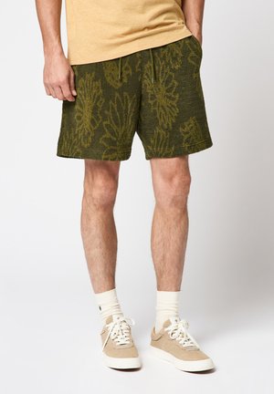 Homme portant un short vert à motifs floraux, des baskets beiges, des chaussettes blanches et un T-shirt beige, debout devant un fond uni.