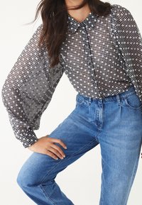 Una persona in una blusa nera e bianca a quadri trasparente e jeans blu posa con una mano sulla vita, mostrando un outfit casual.