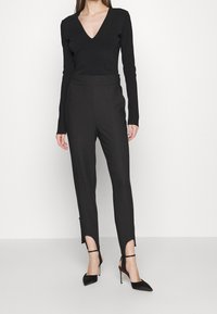 Svart åtsittande långärmad topp med V-urringning, tillsammans med skräddarsydda svarta byxor med utskärningar vid fållen och svarta högklackade skor.