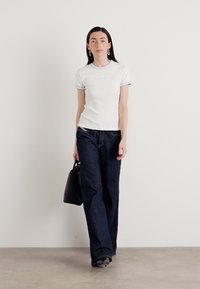 Witte aansluitende t-shirt met blauwe rand, gecombineerd met donkere wijde jeans. Model houdt een zwarte handtas vast en staat voor een neutrale achtergrond.