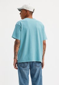 Tricou din bumbac turcoaz, cu mânecă scurtă și croială lejeră. Are un gât rotund și o textură netedă, purtat cu blugi denim albaștri.