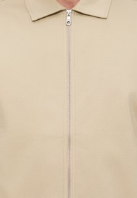 Veste beige à col avec une fermeture éclair argentée centrée, courant verticalement à l'avant.