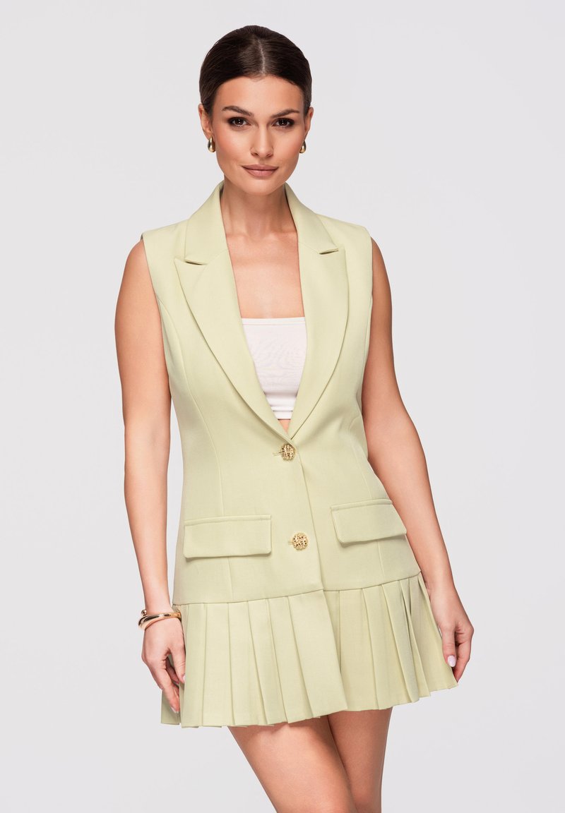 Femme aux cheveux foncés portant une robe blazer jaune clair sans manches avec des boutons dorés et une jupe plissée, posant devant un fond uni.
