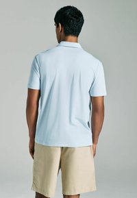 Helles blaues Polohemd mit kurzen Ärmeln, glatter Textur und klassischem Kragen, kombiniert mit beigen Shorts. Rückenansicht des Outfits.