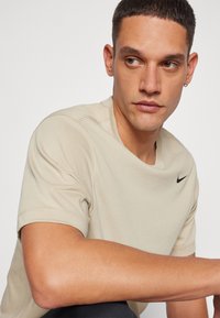 Bež lühikeste varrukatega Nike T-särk, millel on ümmargune kaelus ja musta värvi logo. Sile tekstuur ja korrapärane disain, mis rõhutab õlavarre kontuuri.
