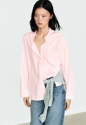 Femme aux cheveux noirs portant une chemise ample rose clair boutonnée, un jean bleu et un pull gris noué autour de la taille.
