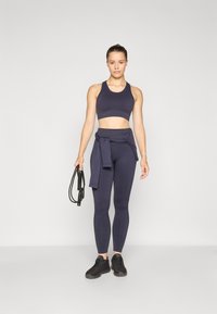 Mørk marineblå sports-bh og høytlivs leggings med teksturert finish. Holder et hoppetau, iført svarte treningssko.