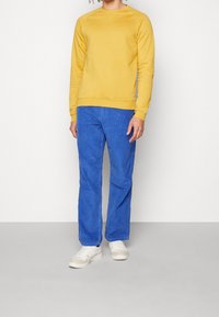 Gul sweatshirt med rund halsringning och ribbade ärmslut i kombination med blå kordbyxor. Kompletteras med vita sneakers med bruna detaljer.