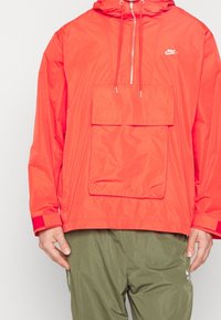 Röd vattentät jacka med halvdragkedja fram, justerbar huva och stor framficka. Har elastiska ärmslut och Nike-logotyp.