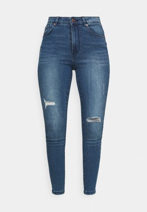 Tamno plave skinny traperice, sa istrošenim detaljima i riječnim rupama na koljenima, standardni dizajn s pet džepova i kopčanjem na gumb.
