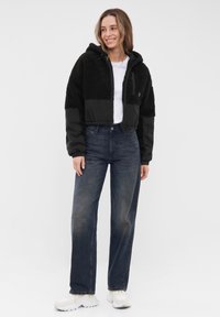 Giacca nera cropped con una texture morbida e cappuccio, abbinata a jeans di denim scuro a vita alta e sneakers bianche. Design semplice e funzionale.