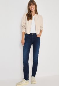 Chaqueta bomber beige con puños elásticos, camiseta blanca, vaqueros azul oscuro y zapatillas de color claro. El atuendo tiene un ajuste casual.