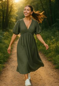 Olijfgroene jurk met korte pofmouwen, V-hals en een getailleerde taille, gecombineerd met witte sneakers, gedragen door een glimlachend persoon op een pad.