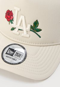 Μπεζ ρυθμιζόμενο καπέλο με κεντητό κόκκινο τριαντάφυλλο και πράσινα φύλλα. Διαθέτει πεταχτά γράμματα "LA" και αυτοκόλλητο λογότυπο New Era στο γείσο.
