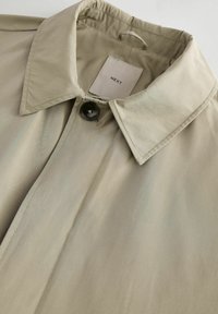 Khaki trenchcoat met een grote kraag, een enkele knoopsluiting en een gladde, licht glanzende textuur. Etiket zichtbaar binnenin de kraag.