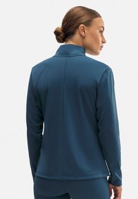 Top deportivo de manga larga en color verde azulado con cuello alto, textura suave y diseño ajustado, que presenta una costura central en la parte posterior y mangas largas.