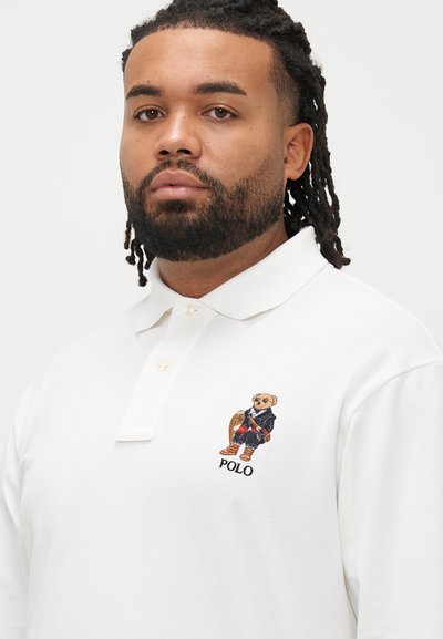 Λευκό πόλο πουκάμισο με κοντά μανίκια, διαθέτει κεντητή αρκούδα που φοράει μπλε ένδυμα και τη λέξη "POLO" κάτω από το σχέδιο.