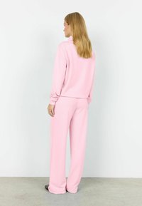 Žena s dlouhými blond vlasy, oblečená v volném světle růžovém svetru a odpovídajících širokých kalhotách, stojící před jednobarevnou bílou zdí.