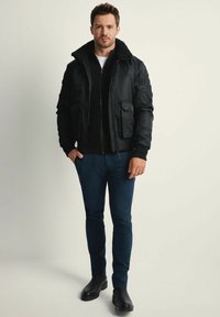 Mężczyzna w czarnej kurtce bomberce z kołnierzem z polaru, granatowych spodniach, czarnych butach, stojący z jedną ręką w kieszeni na neutralnym tle.