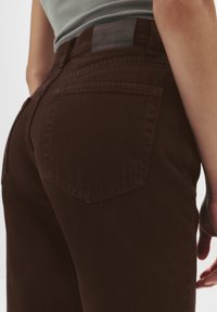 Jeans din denim maro cu o textură netedă, având o etichetă din piele la talie și două buzunare pe spate cu o contur cusut.