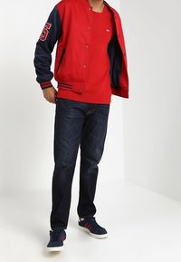 Rote und marineblaue Collegejacke mit Logo- und Nummerndetails, kombiniert mit einem roten Sweater, dunklen Jeans und marineblauen Sneakers. Lässiger sportlicher Stil.