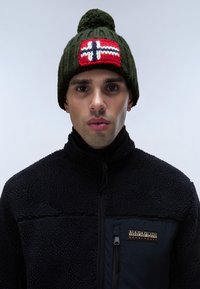 Vihreä neulottu pipo, jossa on pompomi päällä, ja punainen kangas, jossa on valkoinen risti ja sinisiä yksityiskohtia. Pipo on yhdistetty mustan fleece-takin kanssa.