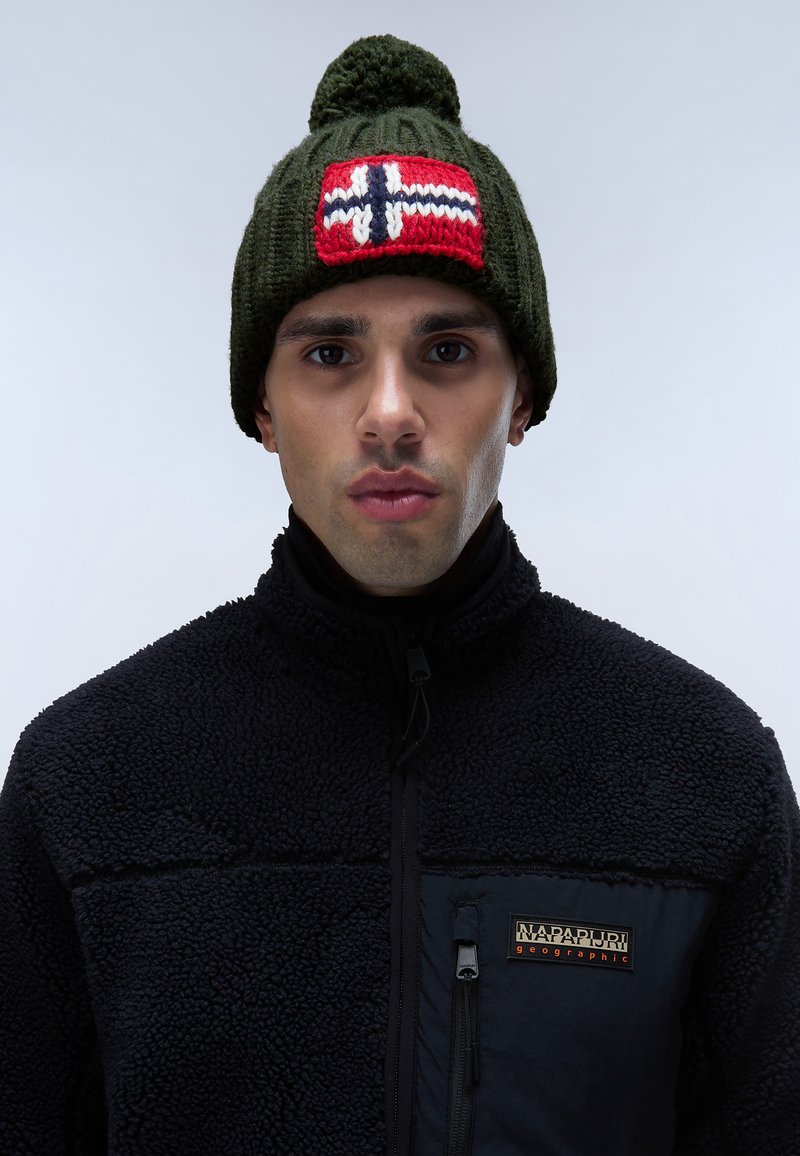 Vihreä neulottu pipo, jossa on pompomi päällä, ja punainen kangas, jossa on valkoinen risti ja sinisiä yksityiskohtia. Pipo on yhdistetty mustan fleece-takin kanssa.