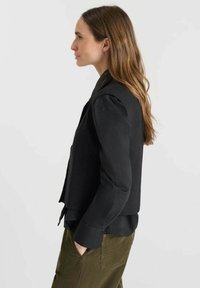 Gilet noir ajusté avec un col structuré, superposé sur une chemise à manches longues foncée. Texture lisse et design ajusté.