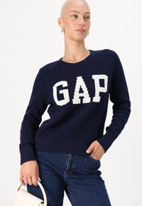 Marineblauwe gebreide trui met een opvallend wit "GAP" logo, een ronde halslijn, geribde mouwen en een losvallende pasvorm. Gecombineerd met donkere denim jeans.