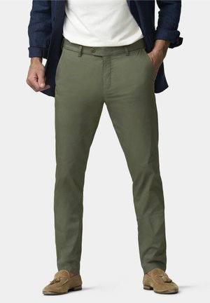 Mann trägt olivgrüne, schmal geschnittene Hose, hellbraune Wildleder-Loafer mit Quasten, weißes Hemd und marineblaue Jacke mit Händen in den Taschen.