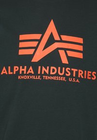 Tricou de culoare verde închis, cu un logo grafic portocaliu ce include o stea și aripi, și textul "ALPHA INDUSTRIES, KNOXVILLE, TENNESSEE, U.S.A."