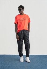 Κόκκινο t-shirt Nike με μαύρο λογότυπο, συνδυασμένο με μαύρο παντελόνι στενής γραμμής. Λευκά αθλητικά παπούτσια, μαύρα γυαλιά ηλίου. Το μοντέλο στέκεται μπροστά σε ουδέτερο φόντο.