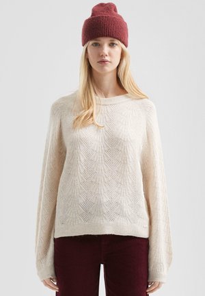 Crème gebreide trui met bladdessin, losse pasvorm, ronde halslijn, en lange mouwen, gecombineerd met een bordeaux ribgebreide beanie.