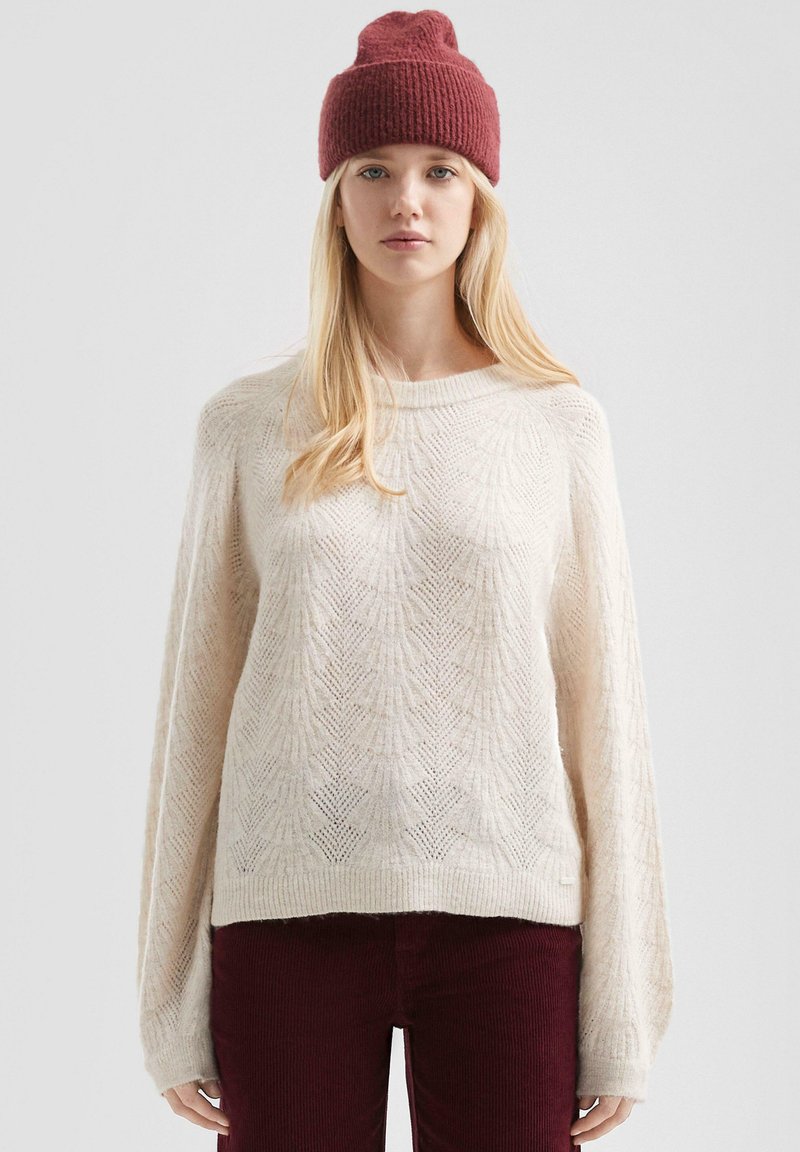 Crème gebreide trui met bladdessin, losse pasvorm, ronde halslijn, en lange mouwen, gecombineerd met een bordeaux ribgebreide beanie.