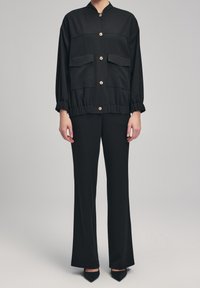 Veste noire à col avec boutons dorés, deux poches avant, taille élastique, et pantalon noir assorti à coupe évasée et texture lisse.