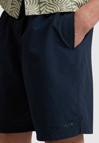 Shorts bleu marine en tissu léger, avec des poches latérales et un logo de marque discret près de l'ourlet. Texture lisse et coupe décontractée.