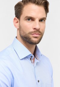 Camicia azzurra chiara con un motivo testurizzato, colletto strutturato e dettagli interni del colletto a contrasto con piccoli punti e accenti marroni.