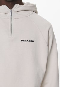 Világosszürke pulóver kapucnis felső, negyed cipzárral és bordázott mandzsettákkal, a bal mellen fekete "PEGADOR" logóval. Puha tapintás, laza dizájn.