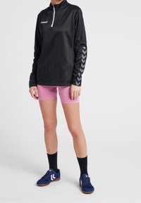 Camiseta atlética de manga larga negra con cremallera de cuarto de vuelta y acentos reflectantes en forma de chevron, combinada con pantalones cortos rosas y zapatillas deportivas azul marino.
