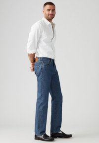 Jeans in denim blu con taglio dritto, abbinati a una camicia bianca con bottoni. Presentano una toppa in pelle sul retro e scarpe eleganti nere.