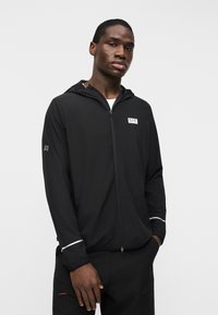 Νέος άνδρας που φοράει μαύρο μπουφάν με κουκούλα και φερμουάρ, με το λογότυπο EA7 στο στήθος και ανακλαστικές ρίγες στα μανίκια, στέκεται με τα χέρια στις τσέπες.