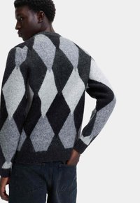 Giovane uomo che indossa un maglione a motivi rombi in nero, grigio e bianco, in piedi con una mano nella tasca posteriore dei jeans scuri.