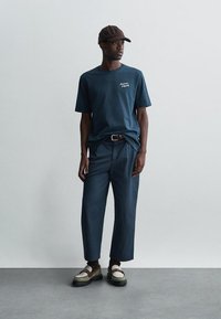 Mann iført mørkeblå t-skjorte, korte mørke bukser, brun cap og beige loafers som står mot en lys, ensfarget vegg.