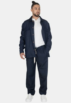 Uomo con capelli raccolti che indossa una polo bianca, una giacca blu scuro abbottonata e rilassata, pantaloni abbinati con coulisse e sneakers bianche.