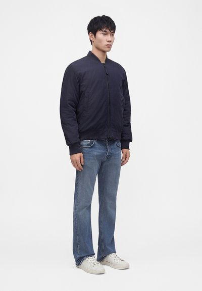 Jeune homme debout portant une veste bomber bleu marine foncé, un jean bleu et des baskets blanches sur un fond blanc uni.