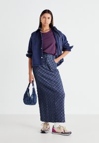Veste en denim bleu foncé, haut violet, jupe longue bleue texturée et basket beige avec des accents roses. Tient un sac à main à motifs bleus.