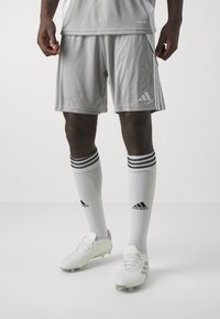 Sivé športové šortky s tromi bielymi pruhmi, kombinované s bielymi ponožkami s čiernym logom a bielymi futbalovými kopačkami.