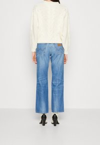 Pull en maille torsadée crème à manches longues, associé à un jean bleu clair à jambes larges avec poches arrière et une étiquette en cuir. Bottines noires à talons.